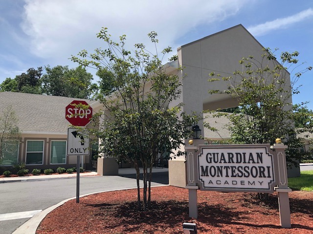 The front of Guardian Montessori Academy 145 Doctors Village Dr., St. Johns FL. 32259. A single story building with a car drive. The building has a covered drive area in the front to provide the ease of loading and unloading of children. There are a few young trees in the picture. And a Sign saying Guardian Montessori Academy.